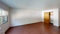 This single-floor unit offers a straightforward, functional layout with consistent hardwood-style flooring running throughout the living areas. The floor plan, as seen in the 3D overview, includes a living space, a galley-style kitchen, at least one additional room, and a full bathroom, all connected in a practical arrangement.

The living area is generously sized, featuring white walls, wood-toned baseboards, and large windows that allow natural light to fill the space. A wall-mounted air conditioning unit and baseboard heating provide year-round climate control. Views from the windows look out onto a green, tree-lined exterior setting.

The galley kitchen is efficiently designed, featuring stainless steel appliances including a coil-burner electric range and a refrigerator, wood-toned cabinetry offering ample storage, and a range hood above the stove. Counter space lines both sides of the narrow kitchen corridor.

A secondary room, also with hardwood-style flooring and a warm wood-toned door, offers a clean, versatile space. The full bathroom includes tile wainscoting, mosaic tile flooring, a white vanity with cabinet storage, a mirrored medicine cabinet with built-in lighting, and a bathtub with a curtained shower. Overall, the unit presents a clean and functional living environment.