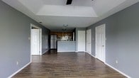 This single-floor space presents an open-concept layout that connects the living area directly to the kitchen, creating a functional and cohesive flow throughout. The flooring consists of rich, dark wood-look tile planks that run uniformly across the entire space, providing both durability and visual warmth against the cool gray walls.

The living area benefits from a tray ceiling detail, which adds architectural interest and a sense of height to the room. A ceiling fan is mounted centrally within the tray, ensuring comfortable airflow throughout the space.

The kitchen is positioned at the far end of the open floor plan and features dark wood cabinetry, a stainless steel refrigerator, and a stove with an over-the-range microwave. A tile backsplash adds texture and detail to the cooking area, while a half-wall peninsula subtly defines the kitchen from the living space without interrupting the open feel.

Multiple white panel doors are visible along the walls, providing access to additional rooms or storage. White baseboards and trim throughout offer a clean contrast to the gray walls, contributing to a crisp, well-defined interior aesthetic.