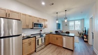 This well-appointed single-floor residence showcases a thoughtfully designed open-concept layout with modern finishes throughout. The kitchen serves as a central highlight, featuring light maple cabinetry with dark hardware, sleek black countertops, a white subway tile backsplash, and a full suite of stainless steel appliances including a refrigerator, range, built-in microwave, and dishwasher. A peninsula with an overhanging dark countertop and bar seating is illuminated by two stylish geometric pendant lights, making it ideal for casual dining or entertaining. Light wood-style flooring flows consistently throughout the space, contributing to a cohesive and contemporary aesthetic. The bedroom is a calm retreat with neutral tones, natural light from multiple windows, and a built-in closet with shelving. A flexible secondary room functions effectively as both a living area and a home office, featuring a window with street views and ample space for multiple furniture configurations. The bathroom is finished with large-format slate-toned floor tiles, a rich wood vanity, white quartz countertop, brushed nickel fixtures, and a frameless mirror. Recessed lighting and clean white walls throughout the space create a bright, airy atmosphere.