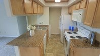 This single-floor residence offers a well-organized and functional living environment. The kitchen is a standout feature, designed in a classic galley layout that maximizes efficiency and storage. Warm honey-toned wood cabinetry lines both the upper and lower sections of the kitchen, providing generous storage space throughout. The countertops feature a granite-look laminate surface that adds a touch of warmth and durability to the workspace. A stainless steel sink is conveniently positioned along one counter run, complemented by a built-in dishwasher beneath. On the opposing wall, a white electric coil-top range and oven sit beneath an over-the-range microwave, while a full-size refrigerator anchors the end of the run. A recessed ceiling detail above the kitchen adds subtle architectural interest and helps define the cooking space. The flooring consists of neutral stone-look tile, which is both practical and easy to maintain. Soft, neutral wall tones throughout create a calm and inviting ambiance. A doorway at the far end of the kitchen connects seamlessly to the rest of the home, supporting a smooth and logical flow through the single-level floor plan.