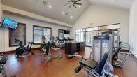 Located at 5252 Riggins Run in Dublin, Ohio, this single-floor fitness facility offers a comprehensive and thoughtfully designed wellness environment. The centerpiece is a generously sized exercise room with soaring vaulted ceilings, rich hardwood-style flooring, and an abundance of natural light streaming through large, dark-trimmed windows. The space is equipped with a wide array of Life Fitness cardio machines — including treadmills, ellipticals, and rowing machines — as well as strength-training equipment and spin bikes. Full-length mirrors and a wall-mounted TV enhance the workout experience, while a ceiling fan overhead adds comfort. A dedicated studio room features wall-mounted TRX suspension systems, balance accessories, stability balls, and open floor space ideal for stretching or group classes. This room also benefits from ample natural light and polished hardwood floors. A standout feature is a dramatic multi-story climbing wall, visible through glass partitions and rising through a soaring atrium. The hallway entry area offers carpeted flooring and large windows with outdoor views of the surrounding neighborhood. The bathroom is finished with tasteful botanical-print wallpaper, tile flooring, a woven rug, and a tanning booth, creating a spa-like transition space for guests.