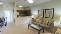This two-floor residence offers a thoughtfully designed open-concept layout that connects the living area and kitchen seamlessly. The living room is finished with plush wall-to-wall carpeting and painted in a soft sage green, creating a calm and welcoming atmosphere. A ceiling fan with a light fixture overhead provides both airflow and ambient lighting, while decorative framed botanical artwork adorns the walls, adding a refined touch to the space.

The living area accommodates comfortable seating and is anchored by a glass-top coffee table with a metal frame, complemented by a matching side table and a table lamp that adds warm accent lighting. The open layout allows for easy sightlines into the kitchen, which is separated by a raised breakfast bar countertop — ideal for casual dining or entertaining.

The kitchen features rich dark wood cabinetry, stainless steel appliances including a built-in microwave and refrigerator, and dark countertops that contrast neatly with the lighter walls. A hallway visible from the main living area suggests additional rooms and access to the second floor, reinforcing the spacious and functional nature of this multi-level residence.