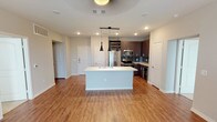 This thoughtfully designed single-floor residence blends contemporary style with everyday functionality. The heart of the unit is a sleek, open-concept kitchen featuring dark espresso cabinetry, a tiled backsplash in cool blue-gray tones, stainless steel appliances including a refrigerator, range, microwave, and wine cooler, a built-in wine rack, and a central island with a white countertop and integrated sink. Pendant lighting above the island adds a modern touch, while warm wood-look flooring flows throughout the kitchen and living areas. The living space is bright and open, with a ceiling fan and French doors that invite natural light and provide access to an outdoor balcony area. Multiple bedrooms offer plush carpeting, ceiling fans, and windows with views of surrounding greenery, creating a comfortable and tranquil atmosphere. Two bathrooms serve the unit: one features a tiled walk-in shower and direct access to a walk-in closet with built-in shelving, while the second includes a soaking tub and shower combination with dark tile surround, granite countertops, and dark wood cabinetry. The neutral color palette throughout creates a clean, cohesive aesthetic well-suited for modern living.