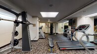 This fitness center is a fully appointed exercise facility designed to accommodate a wide range of workout routines. The space is laid out across a single level, with patterned carpet tiles covering much of the floor and protective rubber matting in designated strength training zones. Overhead, a large skylight panel and recessed lighting provide consistent, bright illumination throughout the room.

The cardio area is anchored by multiple Star Trac treadmills, offering ample space for users to exercise simultaneously. A multi-station cable machine by Hoist and a Body Solid functional trainer provide versatile options for resistance and strength training, while a TKO cable machine adds further variety. A flat weight bench with a barbell setup is centrally positioned for free-weight exercises. A well-stocked dumbbell rack holds a full range of weights, neatly organized for easy access.

Floor-to-ceiling mirrors along one wall visually expand the space and allow users to monitor their form during workouts. A stability ball and additional accessories are available for functional training. An instructional fitness chart is posted on the wall for reference. The facility also includes a wall-mounted television and leads to a secondary corridor area with additional equipment and an emergency exit, ensuring both convenience and safety throughout the space.
