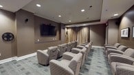 This dedicated screening room delivers an immersive, cinema-inspired experience within a refined and thoughtfully designed space. The room is arranged with multiple rows of plush, upholstered lounge chairs in a neutral patterned fabric, all oriented toward a large wall-mounted flat-screen display positioned at the front of the room. Recessed ceiling lighting provides a warm, ambient glow, while wall-mounted speakers support a quality surround sound setup. The walls are finished in deep, earthy brown tones that enhance the theater atmosphere, and crisp white crown molding adds a touch of elegance to the overall aesthetic. Floor-to-ceiling draping in a rich gold and taupe fabric lines one wall, contributing to both the acoustics and the upscale ambiance of the room. A collection of framed contemporary artwork adorns one side wall, adding a gallery-like quality to the space. At the rear, a small credenza with a microwave offers a convenient refreshment station. Patterned carpet in muted tones of gray and blue covers the floor, completing the polished, cinema-lounge feel. The space is designed to comfortably accommodate a group for screenings, presentations, or private gatherings, combining functionality with a sophisticated aesthetic throughout.