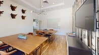 Located at 504 E Ninth St in Tucson, Arizona, this single-floor space offers a thoughtfully designed layout comprising a dining room and a private office. The dining area serves as a versatile meeting and collaboration space, featuring warm hardwood-style flooring, crisp white walls, and a striking brass sputnik chandelier that anchors the room. Multiple wooden tables with mixed seating provide ample space for gatherings, while a large whiteboard mounted on the wall supports presentations and brainstorming sessions. A window with a plaid roman shade invites natural light and views of the surrounding neighborhood. Decorative bull head wall mounts add a bold, eclectic touch to the room's ambiance. The adjoining office is equally well-considered, with built-in and wall-mounted desks lining the perimeter, ergonomic seating, and two distinctive industrial-style dome pendant lights with teal interiors. A geometric ceiling accent and patterned roman shade add visual interest, while a wall-mounted shelving unit provides practical storage. A glass-paneled door connects the spaces, maintaining an open, airy feel throughout. Together, the rooms create a functional yet character-rich environment ideal for professional work and collaborative meetings.