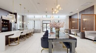 Located on Southern Boulevard in West Palm Beach, Florida, this single-floor space presents a meticulously designed interior that blends modern elegance with warm, luxurious detailing. The open layout is anchored by polished marble-look porcelain tile flooring that flows seamlessly throughout, reflecting abundant natural light streaming in through large windows dressed in sheer white floor-to-ceiling curtains.

The main gathering area features a striking dark marble-top table surrounded by deep navy velvet barrel chairs, illuminated by a sculptural brass multi-globe chandelier overhead. Adjacent, a long white quartz bar counter is lined with white and gold stools beneath a row of clear glass globe pendant lights, complemented by a dark wood built-in shelving unit with a wet bar and stainless steel refrigerator. A secondary seating area showcases cream upholstered chairs and white dining chairs paired with gold-framed tables.

A decorative warm-toned lattice screen partition defines a lounge nook furnished with a plush neutral sofa and accent chairs. Tall tropical palms in brushed gold planters add organic texture near the glass entry doors, which open to an outdoor terrace. Full-length mirrors and warm wood wall panels further enhance the refined, welcoming ambiance throughout.