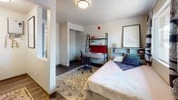 Located at 1555 NE Merman Drive in Pullman, Washington, this single-floor unit offers a compact and efficiently designed living space suited for student or individual living. The main room serves as a combined bedroom and living area, finished with dark hardwood-style flooring, a textured ceiling, and neutral-toned walls that create a clean, understated atmosphere. A large window allows generous natural light to fill the room while offering views of the surrounding neighborhood and adjacent building exteriors. The space accommodates a sleeping area alongside a dedicated study zone, supported by a built-in bookcase headboard and a freestanding shelving unit with a desk workspace, making it well-suited for academic life. A pass-through opening in the wall reveals a kitchenette nook outfitted with a sink, cabinetry, and patterned tile backsplash, providing essential food preparation convenience without occupying the main living area. The private bathroom features wood-style flooring, a wall-mounted pedestal sink with a modern matte black faucet, a large mirror, and a walk-in shower with a curtain enclosure. The entry door opens directly into the main living space, contributing to an efficient, single-level layout. Overall, the unit presents a practical and comfortable environment with thoughtful use of space throughout.
