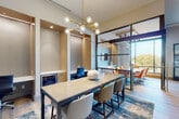 This thoughtfully designed co-working space combines open collaboration with focused, private work areas, all unified by a cohesive contemporary aesthetic. At the heart of the space sits a central communal table topped in a neutral finish and surrounded by upholstered chairs, anchored by a bold blue abstract area rug and illuminated by a striking gold Sputnik chandelier that serves as the room's focal point.

Along one wall, a series of recessed individual workstations are built into warm, illuminated alcoves, each equipped with a built-in desk surface, a dedicated chair, and under-desk storage — ideal for heads-down, focused work. A separate private office nook features a wall-mounted desk, a textured accent wall, and brass bar lighting for a refined, distraction-free environment.

A glass-partitioned conference room with dark metal framing occupies one side of the space, offering semi-private meeting capability while maintaining visual connectivity to the rest of the area. This room is furnished with a dedicated conference table and chairs and benefits from floor-to-ceiling windows that flood the space with natural light and provide elevated outdoor views of a tree-lined landscape. Light wood-tone flooring flows throughout, complementing the warm neutral palette and modern brass fixtures that lend a polished, professional ambiance to the entire space.