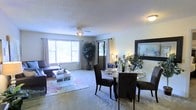 This single-story residence is thoughtfully designed to maximize space and flow across its layout. The floor plan reveals a well-organized arrangement of living areas connected through an open-concept design, allowing natural movement between rooms.

The combined living and dining area serves as the heart of the home. Large windows along the exterior wall flood the space with abundant natural light, creating a bright and welcoming atmosphere. The living area features plush carpeting, complemented by a decorative area rug, and is furnished with comfortable seating centered around a glass coffee table. A ceiling fan overhead adds both functionality and a finishing touch to the room.

The dining area transitions seamlessly from the living space, featuring a round glass-top table surrounded by dark upholstered chairs, ideal for both casual meals and entertaining. Tasteful décor, including indoor plants and accent lighting, adds warmth throughout.

The kitchen is positioned centrally within the floor plan, offering convenient access to the surrounding living areas. Additional rooms, including a bedroom and bathroom, are arranged along the perimeter, providing a practical separation between private and communal spaces. The overall ambiance is warm, light-filled, and inviting.