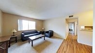 This single-floor residence offers a practical and comfortable layout designed for everyday living. The living room serves as the central gathering space, featuring a combination of plush carpeting and hardwood flooring that transitions into the hallway beyond. A large window along the front wall allows natural light to fill the room, creating a bright and open atmosphere. The textured ceiling adds a classic residential character to the space. Built-in HVAC venting ensures year-round climate control throughout. The living area provides ample room for seating and a central coffee table, making it a functional space for relaxing or entertaining. A hallway extending from the living room leads to additional areas of the home, including what appears to be a bathroom visible in the background. The warm, neutral wall tones throughout lend a cohesive and understated aesthetic to the interior. The hardwood flooring in the corridor adds a contrasting texture to the carpeted main living area, highlighting the thoughtful material choices across the space.