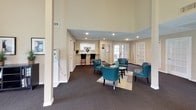 This single-story space is thoughtfully designed to accommodate both collaborative and private work environments. The reception and lounge area greets visitors with dramatically high ceilings, recessed lighting, and plush dark carpeting. Teal upholstered chairs are arranged around a central coffee table atop a neutral area rug, creating an inviting waiting area. Adjacent to the lounge, a compact kitchenette with white cabinetry and counter seating provides convenient utility. French doors along the back wall allow natural light to filter into the space.

The private office areas feature vaulted ceilings accented by ceiling fans and track lighting, with walls painted in a warm sage green and neutral tan combination. Large windows with horizontal blinds line multiple walls, offering generous views of the surrounding landscape. Each office is furnished with a rich mahogany desk and leather chairs, conveying a professional atmosphere. A secondary workspace includes additional desk configurations, cabinetry with decorative accents, and wall art, completing the functional and polished character of this well-appointed space.