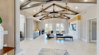 This single-story community clubhouse in Kingsland, Georgia offers a thoughtfully designed gathering space that blends modern elegance with warm, inviting details. The centerpiece of the interior is a dramatic vaulted ceiling adorned with exposed rustic wood beams and a series of striking geometric pendant chandeliers that cast a warm glow throughout the open layout. Light-toned large-format tile flooring in a herringbone pattern runs throughout, enhancing the sense of spaciousness and brightness.

The grand entry foyer features rich wood-paneled walls, decorative art, classic column accents, and a bold set of multi-pane wood-framed glass doors that welcome visitors with an immediate sense of arrival. The expansive main lounge area is furnished with comfortable neutral sofas, armchairs, and a sleek media wall with a large flat-screen TV framed by board-and-batten detailing and warm wood paneling above.

A built-in wet bar with soft blue cabinetry, stone countertops, a beverage refrigerator, and a patterned tile accent wall serves as a stylish refreshment station. Adjacent to the main lounge, a dedicated business office provides a carpeted, quiet workspace equipped with desks and office equipment. French doors along the rear wall open to an outdoor area with direct views of the community pool, seamlessly blending indoor and outdoor living.