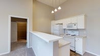 Located at 2828 Marblevista Blvd in Columbus, Ohio, this single-floor residence offers a well-designed layout that balances functionality with an open, airy feel. The kitchen is a standout feature, showcasing dramatic vaulted ceilings that lend a sense of spaciousness well beyond its footprint. White cabinetry lines the walls, providing ample storage, while white appliances — including a refrigerator, electric range, microwave, and dishwasher — offer a clean, cohesive look. A white peninsula counter extends into the living area, creating a natural divide between the kitchen and the adjoining spaces while also serving as a convenient breakfast bar or prep area. Pendant lighting hangs above, adding warmth and focused illumination to the kitchen zone. The flooring transitions from tile in the kitchen to carpet in the living and bedroom areas, offering both practicality and comfort underfoot. A walk-in closet is visible through a doorway, suggesting well-appointed bedroom storage. Neutral wall tones throughout give the space a versatile, move-in-ready feel. Overall, the unit presents a practical and inviting atmosphere suited for everyday living.