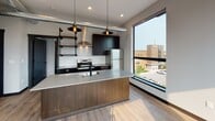 Located in the vibrant Bridge Street corridor of Grand Rapids, Michigan, this single-floor unit showcases a thoughtfully designed modern interior with clean lines and high-quality finishes throughout. The open-concept kitchen serves as a centerpiece, featuring a large island with a quartz countertop, an undermount sink with a matte black faucet, dark cabinetry, stainless steel appliances, a subway tile backsplash, and exposed industrial-style open shelving. Overhead, pendant lights with an Edison-bulb aesthetic add warmth and character, while an exposed ductwork detail reinforces the urban industrial aesthetic. Light wood-tone luxury vinyl flooring flows consistently throughout the space, complemented by dark baseboards that provide sharp contrast. The living area benefits from an abundance of natural light pouring through oversized black-framed windows, offering elevated views of the surrounding neighborhood and mature tree canopy. The bedroom is a calm, light-filled retreat with a tall window and neutral walls. The bathroom is finished with light flooring, a full tub-shower combination with matte black hardware, a dark wood vanity, and clean white surfaces. The overall ambiance is contemporary and urban, balancing industrial-chic elements with bright, livable comfort.