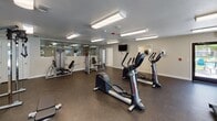 This single-floor fitness center is thoughtfully designed to accommodate a wide range of workout needs. The main exercise area is spacious and well-lit, featuring bright overhead fluorescent lighting and durable rubber flooring throughout. Large windows along one wall offer views of an outdoor pool and patio area, filling the space with natural light and providing a motivating backdrop during workouts.

The cardio zone is well-stocked with multiple treadmills, elliptical trainers, a stair climber, and a rowing machine, offering ample options for aerobic exercise. A dedicated strength training section includes a multi-station cable machine, weight benches, and a full rack of dumbbells, catering to users at all fitness levels. Wall-mounted mirrors are strategically placed to allow exercisers to monitor their form.

A separate cycling room provides a more focused environment for spin sessions, equipped with both a spin bike and an upright stationary bike. A wall-mounted television and a smaller display screen are positioned for easy viewing during rides. This room also features a large glass partition that connects visually to the main gym area.

Throughout the facility, the neutral color palette, textured ceilings, and clean layout contribute to a functional and inviting atmosphere suited for consistent, focused training.
