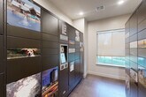 Located at 14698 Briar Forest Drive in Houston, Texas, this dedicated package concierge room is a compact, single-floor amenity space designed to streamline parcel delivery and retrieval. The room is organized around a central corridor flanked on both sides by tall, dark-finished Package Concierge smart locker units. These multi-tiered lockers feature an integrated digital touchscreen interface, allowing residents to securely access their deliveries at any time. The locker doors are adorned with colorful photographic panels showcasing local Houston imagery, adding a visually engaging element to the functional space. The room is finished with neutral, light-toned walls and large-format tile flooring in a warm gray hue, contributing to a clean and modern aesthetic. Natural light filters in through a roller shade-equipped window, brightening the space alongside recessed ceiling lighting. A paneled interior door provides access to an adjacent area, and a clearly marked exit door at the end of the corridor ensures safe egress. The overall design is practical and well-organized, reflecting a thoughtful approach to modern residential amenity spaces.