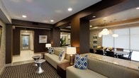 This refined single-floor space makes a striking first impression with its bold design sensibility and thoughtful layout. The living area is anchored by deep chocolate-brown accent walls that create a warm, upscale ambiance, contrasted beautifully by neutral-toned sofas dressed with navy and white geometric-patterned throw pillows. Recessed lighting and stylish table lamps provide layered illumination throughout, enhancing the inviting atmosphere. A patterned area rug grounds the seating arrangement, adding texture and visual depth to the space. A large architectural cutout opening connects the living area to the adjacent dining space, creating a seamless open-concept flow. The dining area features a sleek, dark dining table surrounded by coordinating chairs, illuminated by a pair of elegant globe pendant lights that add a touch of glamour overhead. A granite-topped counter or bar surface bridges the transition between zones, offering both functional and aesthetic value. Framed wall art and curated décor accents are thoughtfully placed throughout, contributing to a cohesive and polished interior. The overall design balances contemporary style with comfortable livability, making this a well-appointed space suited for both entertaining and everyday use.