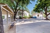 Located at 815 Pomona Ave in Chico, California, this single-level outdoor common area serves as a central gathering space within a multi-building residential community. The grounds are defined by a generous paved courtyard that doubles as a full basketball court, complete with a hoop and clearly marked lines, offering residents an accessible recreational amenity. Mature, broad-canopy trees are distributed throughout the space, casting ample shade across the courtyard and creating a cool, comfortable environment even on sunny California days. A slatted wooden pergola structure along the perimeter provides additional shade and houses seating benches, making it a functional spot for relaxation and socializing. The surrounding buildings feature a stucco exterior in a neutral earth tone, with neatly trimmed shrubs and landscaped beds lining the building facades and walkways. Concrete pathways connect the various sections of the property, ensuring ease of movement throughout the grounds. On-site parking is conveniently accessible within the complex. The overall ambiance is casual and community-oriented, with the combination of open paved areas, mature tree coverage, and dedicated recreational space creating an inviting outdoor environment suited to everyday residential use in the Central Valley climate.