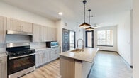 This single-floor residence showcases a contemporary design with a cohesive neutral palette and durable light-toned wood-style flooring running throughout the main living areas. The open-concept kitchen serves as the centerpiece of the space, anchored by a large granite-topped island with an undermount sink and a brushed gold pull-down faucet, illuminated by a pair of stylish pendant lights with bronze-toned shades. Light wood-grain cabinetry lines the walls, complemented by a subway tile backsplash and stainless steel appliances, including a refrigerator, range, and dishwasher. A set of panel doors conceals in-unit washer and dryer connections, adding practical convenience. The kitchen flows seamlessly into the living area, which receives ample natural light through large windows and is equipped with a ceiling fan. Two bathrooms serve the residence; one features a soaking tub with a marble-style tile surround, while the other includes a granite vanity with an undermount sink and a large mirror. Both bathrooms are fitted with accessibility grab bars and light wood-tone cabinetry. The carpeted bedrooms offer a quiet, comfortable retreat, each with a window admitting natural light. Walk-in closets with wire shelving provide organized storage.