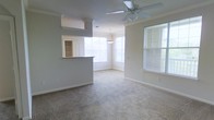 This single-floor space offers an open and airy layout, thoughtfully designed to maximize functionality and comfort. The living area features soft carpeting underfoot and is equipped with a ceiling fan, providing both comfort and airflow. Neutral-toned walls complement the light-filled interior, creating a calm and welcoming ambiance throughout.

A peninsula-style pass-through counter connects the kitchen to the main living space, offering a practical divide while maintaining the open feel of the floor plan. Built-in shelving is visible through the pass-through window, adding convenient storage to the kitchen area. A wall-mounted unit is also present in the living space, adding to the room's utility.

Adjacent to the living area, a dining space is highlighted by a pendant light fixture and is surrounded by a bay of windows that flood the area with natural light. An additional window on the side wall further enhances brightness and ventilation throughout the space. White blinds on all windows offer privacy while still allowing light to filter in. The overall design is clean, neutral, and move-in ready, offering a versatile canvas for a variety of interior styles.