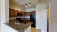 This single-story space centers around a functional and well-appointed kitchen that serves as its defining feature. The kitchen showcases rich, warm-toned wood cabinetry that extends along multiple walls, providing generous storage both above and below the countertops. The dark granite countertops add a touch of elegance and contrast against the lighter cabinetry, running along the perimeter and forming an L-shaped layout that maximizes workspace.

A suite of black appliances, including a refrigerator, electric range, and built-in microwave, lends a cohesive, modern aesthetic to the kitchen. The double-basin sink is conveniently situated within the extended counter space, enhancing the kitchen's practicality. Light neutral tile flooring runs throughout, contributing to a clean and bright atmosphere.

Overhead, a track lighting system with adjustable spotlights ensures the kitchen is well-lit and adaptable to various needs. A set of bi-fold doors nearby suggests additional storage or utility space. The neutral wall tones keep the overall ambiance open and inviting, complementing the warm wood and dark stone finishes throughout.