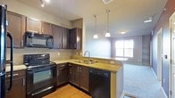This single-floor residence offers a thoughtfully designed open-concept layout that seamlessly connects the kitchen and living areas, creating a spacious and functional living environment. The kitchen is a standout feature, showcasing rich dark espresso cabinetry that contrasts beautifully with the warm granite countertops. Black appliances, including a range, microwave, and dishwasher, lend a sleek, cohesive aesthetic throughout the space. A double-basin stainless steel sink is positioned at the peninsula, which also serves as a natural divider between the kitchen and the adjoining living area. Elegant pendant lights hang above the peninsula, adding a stylish focal point and enhancing the open feel of the space. The kitchen transitions smoothly into the living area, where plush carpeting provides a warm and comfortable atmosphere. A window along the far wall allows natural light to flow into the living space, brightening the interior and offering a view of the outdoors. Hardwood-style flooring in the kitchen adds warmth and durability underfoot. The overall ambiance of the residence is modern and inviting, with carefully chosen finishes that balance style and everyday practicality.