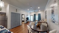 This single-floor unit offers a thoughtfully designed open-concept layout that seamlessly connects the kitchen, dining, and living areas. The space is finished with warm hardwood-style flooring throughout the main living areas and crown molding that adds a polished architectural touch. The kitchen is well-equipped with dark gray cabinetry, stainless steel appliances, subway tile backsplash, light countertops, and pendant lighting above a breakfast bar with seating — ideal for casual dining or entertaining. Adjacent to the kitchen, a round dining table provides a defined dining area, while the living room features a tufted sofa, decorative mirror wall art, and plantation-shuttered windows dressed with teal drapes that allow for natural light and privacy. A ceiling fan with recessed lighting keeps the main living space bright and comfortable. The bedroom is a calm retreat with plush carpeting, layered window treatments, and ample space for furnishings. The bathroom includes a dark vanity with a stone-topped sink, overhead cabinetry, and direct access to the dedicated in-unit laundry room, which is equipped with a full-size washer and dryer and additional storage cabinetry above.