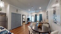 This single-floor unit offers a thoughtfully designed open-concept layout that seamlessly connects the kitchen, dining, and living areas. The space is finished with warm hardwood-style flooring throughout the main living areas and crown molding that adds a polished architectural touch. The kitchen is well-equipped with dark gray cabinetry, stainless steel appliances, subway tile backsplash, light countertops, and pendant lighting above a breakfast bar with seating — ideal for casual dining or entertaining. Adjacent to the kitchen, a round dining table provides a defined dining area, while the living room features a tufted sofa, decorative mirror wall art, and plantation-shuttered windows dressed with teal drapes that allow for natural light and privacy. A ceiling fan with recessed lighting keeps the main living space bright and comfortable. The bedroom is a calm retreat with plush carpeting, layered window treatments, and ample space for furnishings. The bathroom includes a dark vanity with a stone-topped sink, overhead cabinetry, and direct access to the dedicated in-unit laundry room, which is equipped with a full-size washer and dryer and additional storage cabinetry above.