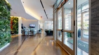 This single-floor lobby and leasing center presents a thoughtfully designed, contemporary space that blends functional amenities with upscale aesthetic details. The entry corridor is anchored by large floor-to-ceiling glass doors and expansive windows that frame views of surrounding greenery and the urban streetscape beyond, flooding the interior with abundant natural light. Textured stacked-stone columns provide a striking architectural contrast against the clean white walls throughout.

Highly polished concrete floors extend across the entire space, reflecting the warm ambient glow of recessed track lighting and cove illumination installed along the ceiling. The open lounge area features stylish circular woven armchairs and a wall-mounted flat-screen television, creating a welcoming, residential atmosphere. Adjacent to the lounge, a glass-enclosed meeting or leasing room is furnished with a distinctive live-edge wooden table surrounded by emerald green upholstered chairs, beneath a cluster of modern pendant lights. A herringbone wood-paneled wall and marble wainscoting add rich texture to this area.

Further within the space, a dedicated mailroom section houses an organized bank of resident mailboxes alongside keypad-controlled access points. A corridor with wood-accented elevator doors and curated wall art connects the lobby to additional building areas, completing a cohesive and polished communal environment.