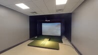 This single-floor space is purpose-built around an indoor golf simulator experience, divided into two distinct functional zones. The simulator bay is the centerpiece, housing a TruGolf system complete with a large projection screen encased in a dark surround enclosure. An artificial turf hitting mat with an elevated tee area is positioned on the floor, providing an authentic feel during play. Overhead, a drop tile ceiling with recessed lighting illuminates the bay, while dark wall treatments minimize glare and enhance the visual immersion of the projected course imagery.

Adjacent to the simulator bay is a control and lounge area finished in neutral gray tones with commercial-grade carpet flooring. This room features a rich dark wood credenza with multiple cabinet doors and gold hardware, supporting a desktop computer used to manage the simulator system. A large flat-screen display is mounted prominently on the wall, ideal for reviewing shot data or showcasing course visuals. A cushioned accent chair offers comfortable seating. A pass-through window connects the two areas, and additional equipment, including what appears to be a secure storage unit, is also present. Drop tile ceilings with flush panel lighting carry throughout both areas, giving the entire suite a clean, professional atmosphere.