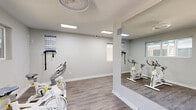 The Ace Fitness Center in Dallas, Texas offers a thoughtfully designed workout space that maximizes functionality within a compact footprint. The interior features a neutral gray-and-white color palette, with light wood-style flooring that runs throughout and bright flush-mounted and recessed lighting that keeps the space well-illuminated at all times.

The facility is organized into distinct workout zones. A dedicated cycling area houses multiple stationary spin bikes, wall-mounted resistance bands, and large floor-to-ceiling mirrors that visually expand the room and allow users to monitor their form. Motivational workout charts are posted on the walls for quick reference.

A long, corridor-style section of the gym serves as the primary cardio and strength training zone, equipped with a treadmill, elliptical cross-trainer, free weights, and a stability ball. Circular pendant lighting fixtures add a modern touch overhead, while black-and-white photographic artwork lends a sophisticated, motivational atmosphere. A gray storage cabinet provides convenient organization for accessories and supplies.

Natural light filters in through multiple windows, and a dedicated entry door offers easy access. Signage for The Ace Fitness Center is prominently displayed near the entrance, establishing the space's identity clearly. The overall ambiance is clean, modern, and energizing — designed to support a focused and effective workout experience.