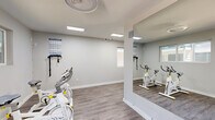 The Ace Fitness Center in Dallas, Texas offers a thoughtfully designed workout space that maximizes functionality within a compact footprint. The interior features a neutral gray-and-white color palette, with light wood-style flooring that runs throughout and bright flush-mounted and recessed lighting that keeps the space well-illuminated at all times.

The facility is organized into distinct workout zones. A dedicated cycling area houses multiple stationary spin bikes, wall-mounted resistance bands, and large floor-to-ceiling mirrors that visually expand the room and allow users to monitor their form. Motivational workout charts are posted on the walls for quick reference.

A long, corridor-style section of the gym serves as the primary cardio and strength training zone, equipped with a treadmill, elliptical cross-trainer, free weights, and a stability ball. Circular pendant lighting fixtures add a modern touch overhead, while black-and-white photographic artwork lends a sophisticated, motivational atmosphere. A gray storage cabinet provides convenient organization for accessories and supplies.

Natural light filters in through multiple windows, and a dedicated entry door offers easy access. Signage for The Ace Fitness Center is prominently displayed near the entrance, establishing the space's identity clearly. The overall ambiance is clean, modern, and energizing — designed to support a focused and effective workout experience.