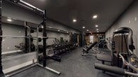 This fitness facility is designed to support a wide range of strength and resistance training routines. The space is finished in a cohesive dark aesthetic, with gray walls, recessed lighting, and durable rubber flooring that provides both safety and functionality throughout. A striking reclaimed wood accent wall serves as a focal point, adding a warm, industrial character that contrasts effectively with the sleek, modern equipment.

The free weights section is well-stocked, featuring an extensive dumbbell rack with a full range of weights, a barbell rack loaded with weight plates, and multiple Matrix adjustable benches to accommodate various exercises. Full-length mirrors line one wall, allowing users to monitor their form during workouts and enhancing the sense of depth in the space.

The resistance and machine training area is equally comprehensive, housing a cable machine system and multiple plate-loaded machines with yellow and black detailing, ideal for targeted muscle group training. Additional benches, including flat and incline options, are distributed throughout the floor to support a variety of lifting routines.

The facility is accessed through a standard door and includes clearly marked exit signage, ensuring safe navigation throughout. The recessed lighting provides consistent, well-distributed illumination across the entire space, creating a focused and motivating environment suited to serious training.