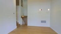 This two-story space is defined by its impressive double-height ceiling, which creates a sense of grandeur and openness in the main living area. The expansive white walls rise dramatically, amplifying the feeling of volume and allowing light to permeate the interior freely. A single pendant light fixture hangs from the elevated ceiling, adding a subtle design element to the otherwise clean and minimalist aesthetic.

The flooring throughout is a warm-toned hardwood, lending a natural and inviting quality to the space. A white-railed staircase leads to the second level, which features an open overlook area, providing visual connectivity between floors. The upper level includes what appears to be a windowed nook or study area, adding architectural interest and functionality to the design.

A standard entry door is visible near the base of the staircase, offering a practical layout transition. A floor-level HVAC vent is integrated seamlessly into the baseboard, maintaining the clean lines of the interior. Overall, the space presents a modern, neutral palette with thoughtful architectural details, making it a versatile and well-structured multi-level environment.