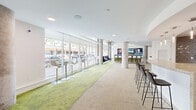 This contemporary single-floor amenity space offers a thoughtfully designed layout that blends social gathering areas with practical amenities. Expansive floor-to-ceiling windows line the exterior wall, flooding the interior with natural light and providing open street views, while exposed concrete columns add an industrial character that contrasts elegantly with the clean white finishes throughout.

The main lounge area features a vibrant combination of green-toned carpet and neutral flooring, creating distinct zones for different activities. A central media bar, upholstered in olive-green fabric and lined with sleek white bar stools, faces a wall-mounted multi-screen display, making it an ideal spot for social gatherings or entertainment. Circular pendant light fixtures hang overhead, adding a refined, modern touch. Adjacent lounge seating with stylish accent chairs offers a more relaxed setting near the windows.

The fully equipped kitchen is tucked within a curved archway and showcases glossy white cabinetry, a richly textured dark brick-style tile backsplash, stone countertops, and stainless steel appliances including a refrigerator and dishwasher. A generous kitchen island with bar seating provides an inviting space for casual dining or conversation. The overall ambiance of the space is bright, modern, and welcoming, designed to foster community and comfort.