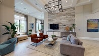 Located at 15616 SW 127th Ave in Miami, FL, this sophisticated single-story community space showcases a seamless open-plan layout designed for comfort and modern living. Light-toned herringbone hardwood flooring runs throughout, lending a refined, cohesive aesthetic to every area.

The expansive main living area is anchored by a dramatic floor-to-ceiling wood-clad accent wall that houses a large flat-screen TV and a sleek white media console. A striking industrial-style rectangular chandelier with exposed Edison bulbs serves as a focal point, illuminating an arrangement of plush seating including a gray sectional, rich cognac leather armchairs, and teal accent chairs. Tray ceilings with recessed lighting add architectural depth overhead.

Adjacent to the main lounge, a dedicated seating nook features teal velvet chairs and connects fluidly to additional intimate lounge spaces. A bright, airy sitting room with soaring ceilings and floor-to-ceiling windows offers sweeping views of lush tropical landscaping and palm trees, inviting abundant natural light.

The dining area is defined by a large reclaimed wood table surrounded by leather chairs, positioned beneath dramatic black-and-gold pendant lights. Expansive floor-to-ceiling windows wrap this space, framing verdant outdoor views and creating a seamless indoor-outdoor connection throughout the entire space.