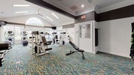 Located at 325 Marymeade Dr in Summerville, SC, this well-appointed fitness center offers a comprehensive workout environment across a single floor. The space is divided into two distinct training zones, each thoughtfully designed to support a wide range of exercise routines. The functional training area features a bold, dark-painted tray ceiling with accent lighting, and a large multi-purpose rig equipped with suspension trainers, gymnastic rings, a heavy bag, and an assortment of medicine balls and free weights. Ceiling fans provide comfortable air circulation throughout. The cardio and strength zone is bright and welcoming, with vaulted ceilings, an arched transom window, and abundant natural light flowing in through large double-hung windows that frame views of the surrounding greenery. This area houses multiple treadmills, an elliptical, and a full suite of selectorized strength machines. Throughout both areas, a vibrant teal and gold geometric-patterned carpet adds a lively, modern aesthetic. Wall-mounted televisions, recessed and linear lighting, and crown molding details contribute to a polished, community-grade facility. The overall ambiance is energetic yet functional, making this fitness center a standout amenity for the Summerville community it serves.