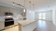 This single-floor residence showcases a contemporary open-concept design that seamlessly connects the kitchen and living areas. The kitchen is a functional centerpiece, featuring gray flat-front cabinetry, a white penny-tile backsplash, and a full suite of stainless steel appliances, including a refrigerator, range, microwave, and dishwasher. A granite-topped island with pendant lighting overhead provides additional prep space and casual seating. Track lighting above the kitchen adds further illumination, while light-toned hardwood flooring extends throughout the main living area, enhancing the sense of openness. Large windows and a glass patio door at the far end of the living space allow generous natural light to fill the room. The bedrooms are carpeted and bright, with sizeable windows offering outdoor views; one bedroom includes a ceiling fan. The bathroom is finished with consistent granite countertops, gray cabinetry with brushed chrome hardware, large-format tile flooring, and a tiled tub-shower surround with a framed mirror above the vanity. The overall aesthetic is clean, modern, and neutral throughout.