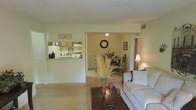 This single-floor residence features an open-concept design that seamlessly connects the main living areas. The living room serves as the central gathering space, furnished with a comfortable sofa and anchored by a dark wood coffee table adorned with decorative candles and a dried floral arrangement, adding a warm, homey touch. Carpeted flooring extends throughout the main areas, contributing to a cozy atmosphere. A pass-through kitchen counter opens directly to the living space, offering a practical and interactive layout. The kitchen features wooden cabinetry and is visible through the pass-through opening, allowing for easy communication between spaces. Adjacent to the living room, the dining area accommodates a bistro-style table with ornate wrought-iron chairs beneath a ceiling light fixture, with a wall clock serving as a decorative focal point. A framed piece of wall art adds character to the dining space. The textured ceiling and neutral wall tones throughout create a clean, understated backdrop. Additional rooms are accessible through a hallway visible beyond the dining area, suggesting further private living spaces within the home.