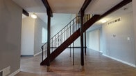 This two-story space is defined by its open, airy main level, where hardwood-style flooring extends throughout the living areas and a distinctive dark wood exposed beam runs across the ceiling, adding architectural character. A bold staircase with ornamental wrought iron railings serves as a central design feature, connecting both floors while contributing a classic, elegant aesthetic to the space.

The main level offers a generous living area illuminated by natural light from vertical blind-covered sliding glass doors and windows that frame views of surrounding greenery. A dedicated entryway area provides a welcoming transition into the home. The kitchen is equipped with wood cabinetry, white appliances including an electric range and dishwasher, and hard-surface flooring, with a window allowing natural light. A half bathroom on the main level features a wood vanity with mirrored cabinet above and tile flooring.

The upper level hosts multiple bedrooms, each carpeted and outfitted with windows offering outdoor views of trees and landscaping. A full bathroom on this level includes a tub-shower combination, a wood vanity with countertop, and tile flooring. Neutral gray tones throughout both floors create a cohesive and calm overall ambiance.