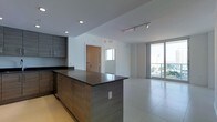 This single-floor space showcases a thoughtfully designed open-concept layout that seamlessly connects the kitchen and living areas. The kitchen is appointed with dark wood-grain cabinetry featuring integrated handles, complemented by a large island with a dark countertop that serves as both a workspace and a visual anchor for the room. Matching upper and lower cabinetry line the adjacent wall, providing ample storage. Recessed lighting and a track lighting system illuminate the kitchen area efficiently.

The living space transitions naturally from the kitchen, featuring large-format light-toned tile flooring that extends throughout, creating a cohesive and clean aesthetic. Floor-to-ceiling glass windows and sliding doors along one wall flood the space with natural light while offering expansive outdoor views and access to a balcony. The bright, airy atmosphere is further enhanced by the white walls and high ceilings. A secondary doorway visible in the background suggests additional rooms beyond the main living area. Overall, the space conveys a contemporary, minimalist character suited for modern living.