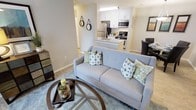 This single-floor space is designed with an open-concept layout that connects the living, kitchen, and dining areas into one cohesive, functional environment. The living area serves as the central hub, featuring light hardwood-style flooring, neutral grey walls, and ample room for comfortable furnishings. A plush area rug anchors the seating space, adding warmth and texture underfoot.

The kitchen is positioned toward the rear of the open floor plan and is well-appointed with white cabinetry, stainless steel appliances — including a refrigerator, microwave, and range — and a light-toned countertop. A peninsula with a sink faces the living area, creating a natural pass-through that enhances the open feel and allows for easy interaction between spaces.

Adjacent to the kitchen, a designated dining area provides a dedicated spot for meals, completing the main living zone. The overall color palette throughout is clean and contemporary, relying on whites, soft greys, and natural wood tones to create a bright, welcoming ambiance. The layout maximizes the use of the available footprint, making the space feel both practical and comfortable.