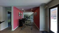 This single-floor fitness space is thoughtfully designed to support a comprehensive workout experience. The room features striking red accent walls that energize the environment, contrasting cleanly with light grey upper walls and a white ceiling. Warm hardwood-style flooring runs throughout, adding a polished, durable surface underfoot.

Recessed ceiling lights provide consistent, even illumination across the space, while a ceiling fan mounted near the entry ensures proper air circulation during intense workouts. A full-length mirrored wall at the far end visually expands the room, creating a sense of greater depth while also serving as a practical training aid for monitoring form.

The space is well-stocked with strength and cardio equipment, including a multi-position adjustable weight bench, a rack of dumbbells, and cardio machines such as ellipticals and treadmills arranged toward the rear. Framed artwork on the walls adds a refined touch, elevating the space beyond a purely utilitarian gym. Overall, the layout is organized and functional, offering an efficient flow between equipment zones within a motivating, well-appointed environment.