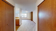 Located at 21428 International Blvd in SeaTac, WA, this single-floor unit offers a thoughtfully arranged layout comprising a living room, kitchen, bathroom, closet, and laundry area. The living room is bright and welcoming, featuring plush carpet flooring and a trio of large windows dressed with vertical blinds that allow ample natural light to fill the space. A pass-through counter with granite-style surfaces connects the living area to the kitchen, which is equipped with warm wood cabinetry, matching countertops, and track lighting overhead. The transition between the kitchen and living areas is marked by a shift from hardwood-style flooring to carpet, creating a clear functional delineation. The bathroom is well-appointed with a full tub-shower combination, a granite-topped vanity with wood cabinetry, a large mirror, and hardwood-style flooring. A standout feature of the unit is the built-in murphy bed housed within a rich wood-paneled cabinet, offering a smart space-saving solution. The closet provides additional storage, while the dedicated laundry area adds everyday convenience. Neutral wall tones and consistent warm wood accents throughout the unit create a cohesive and functional living environment.