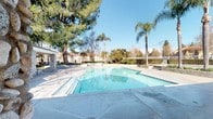 This single-story outdoor recreational facility centers around a generously sized swimming pool with clear, blue-tinted water and a wide surrounding concrete deck ideal for lounging and gathering. The pool area is framed by lush, mature landscaping, including tall shade trees and elegant palm trees that lend a resort-like ambiance under open blue skies.

Adjacent to the pool, a covered pavilion provides shade and shelter, supported by distinctive natural stone columns that add a rustic, architectural character to the space. This covered area offers a comfortable transition between the pool deck and the support facilities, making it well-suited for group use.

The facility includes dedicated restroom facilities housed in a structure with classic horizontal lap siding painted in a soft grey-white finish. Separate men's and women's restrooms are clearly marked and equipped with a mounted fire extinguisher for safety compliance. Keypad entry hardware is installed on the restroom doors, ensuring controlled access.

The perimeter of the pool area is enclosed with fencing, providing privacy and security, while the surrounding greenery contributes to a calm, pleasant atmosphere. The combination of practical amenities, thoughtful landscaping, and durable architectural elements makes this facility a well-equipped and inviting outdoor recreational space.