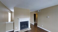 This single-floor residence offers a comfortable and functional layout designed for everyday living. The living area serves as the heart of the home, highlighted by a classic white-mantled fireplace with a glass-front surround, adding warmth and a timeless focal point to the room. Rich, dark hardwood floors extend throughout the main living space, providing a striking contrast against the warm beige walls and crisp white baseboards and trim. The textured ceiling adds a subtle architectural detail overhead, while a ceiling vent ensures comfortable climate control year-round. The open sightlines within the living area create a sense of spaciousness, with a hallway leading to additional rooms beyond, suggesting a well-connected floor plan. Natural light filters into the space, enhancing the inviting atmosphere created by the neutral tones. The transition from hardwood to tile near the fireplace hearth is a practical and visually grounded design choice. Overall, this space presents a clean, well-maintained interior that balances classic residential charm with functional design elements suited for comfortable day-to-day living.