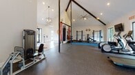 This single-floor fitness center is thoughtfully designed to accommodate a wide range of workout styles. The space is immediately distinguished by its dramatic vaulted ceilings with exposed dark beam accents, creating an open and energizing atmosphere. Recessed lighting and a large ceiling fan ensure the space remains bright and comfortable during workouts.

The cardio zone features multiple treadmills, elliptical machines, and a stationary bike, all positioned near large windows that bring in natural light and offer views of the surrounding greenery. A wall-mounted flat-screen television is conveniently positioned for entertainment during cardio sessions.

The strength and functional training area is anchored by a freestanding multi-purpose rig equipped with a heavy punching bag, battle ropes, suspension trainers, and resistance attachments. Power racks, weight benches, dumbbells, and a weighted sled round out the strength training offerings, all set on durable rubber flooring with a blue padded zone for floor-based exercises.

Motivational phrases, including "Hustle for that Muscle," are displayed prominently on the walls, contributing to an upbeat and encouraging environment. The entry area features wood-look flooring, a hand sanitizer station, and glass doors that allow natural light to flow into the space. The overall design balances functionality with an inviting, modern aesthetic.