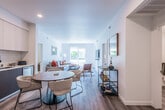 This well-appointed single-floor residence offers a seamless open-concept layout that connects the living, dining, and kitchen areas into one cohesive and functional space. The interior is finished with light wood-look flooring throughout the main areas, crisp white walls, and recessed lighting, creating a bright and airy atmosphere.

The kitchen is a standout feature, equipped with stainless steel appliances including a gas range, over-the-range microwave, and French door refrigerator. Two-tone cabinetry—white upper cabinets paired with charcoal lower cabinets—is complemented by clean white quartz countertops and a classic subway tile backsplash.

The open living area benefits from large windows that invite generous natural light, with views of greenery outside. A dedicated dining space accommodates a round table with seating, while a stylish bar cart area adds a functional accent near the entry corridor.

The residence includes two bedrooms; one features plush carpeting, natural light from a window, and clean white walls, while the other is appointed with designer wall-mounted lighting and a woven headboard. The full bathroom offers a soaking tub with tiled surround, a wide vanity with ample storage, wood-look flooring, and direct access to a spacious walk-in closet.