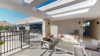 This single-level space centers around a thoughtfully designed outdoor terrace that blends comfort and functionality. The terrace is partially covered by a sleek white pergola structure, allowing natural light to filter through while providing a sense of shelter. A standout architectural feature is a stacked-stone column that anchors the space, adding a warm, textural contrast to the otherwise clean, modern aesthetic of the surrounding stucco walls and light-toned flooring.

The lounge area is furnished with contemporary outdoor seating, including a generously sized sofa and coordinating armchairs in neutral tones, accented with colorful throw pillows. Low-profile coffee and side tables complement the seating arrangement, creating an inviting setting for relaxation or social gatherings. A built-in grill station is positioned nearby, making the terrace well-suited for outdoor dining and entertaining.

A metal railing defines the perimeter of the terrace, with views opening outward to the surrounding area under open blue skies. The overall ambiance is bright and airy, with abundant natural light lending the space a fresh, open-air feel. The design successfully merges indoor-style comfort with outdoor living, resulting in a versatile and welcoming environment.